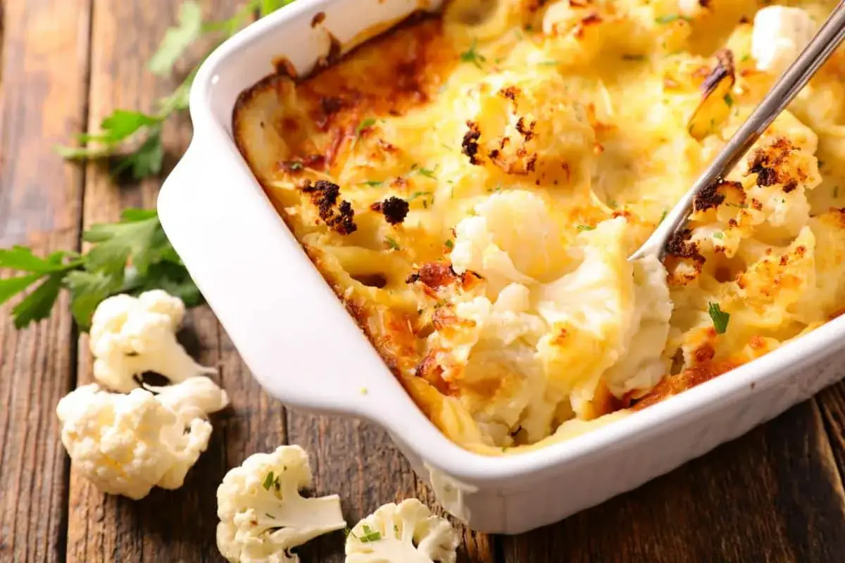 Un gratin de chou-fleur crémeux et doré, servi dans un plat blanc sur fond de bois. Des bouquets de chou-fleur frais sont disposés autour.
