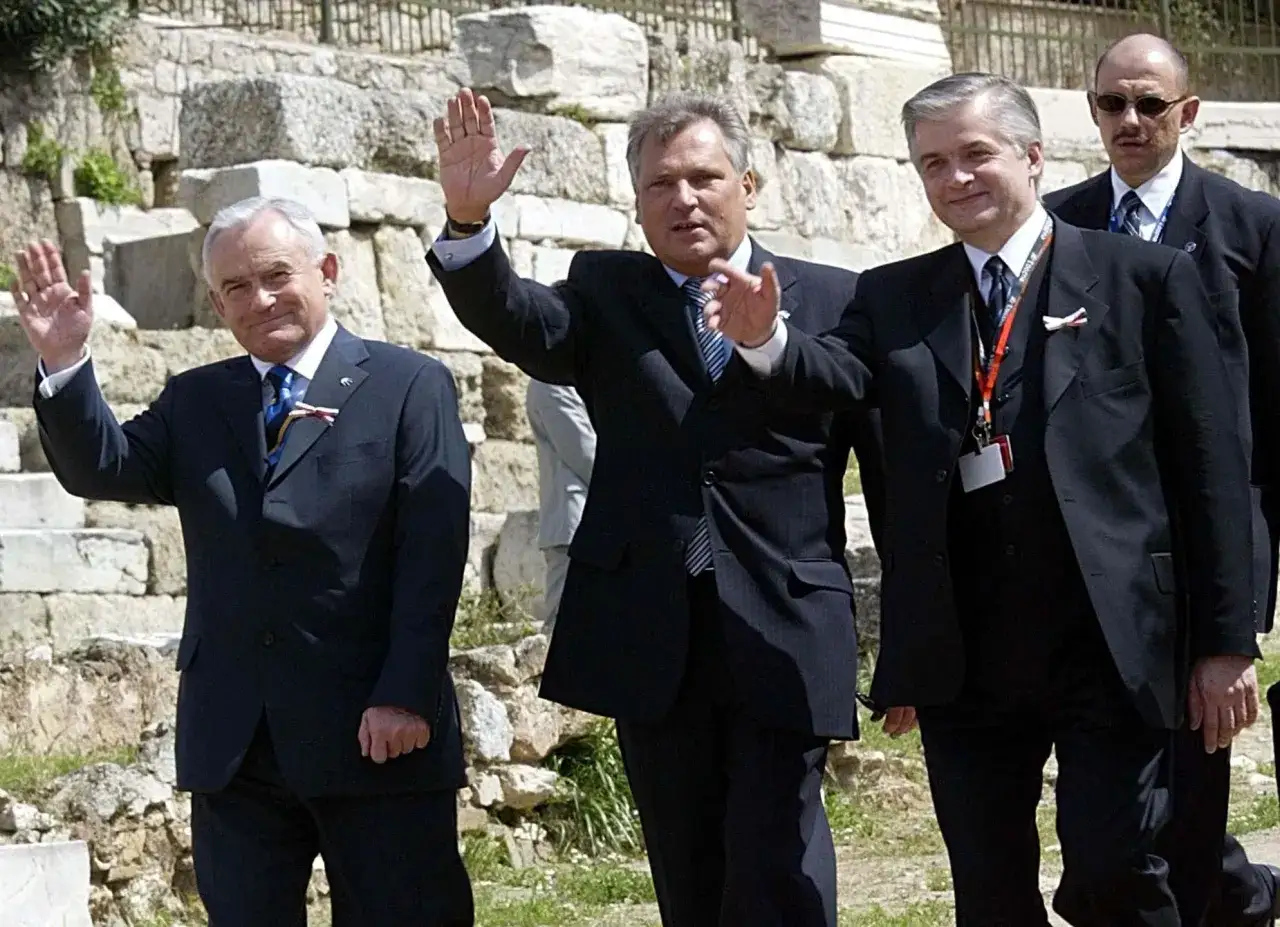 Aleksander Kwaśniewski, prezydentura w Polsce, pozdrawia tłum. Uśmiechnięty, w garniturze, na tle starożytnych ruin.