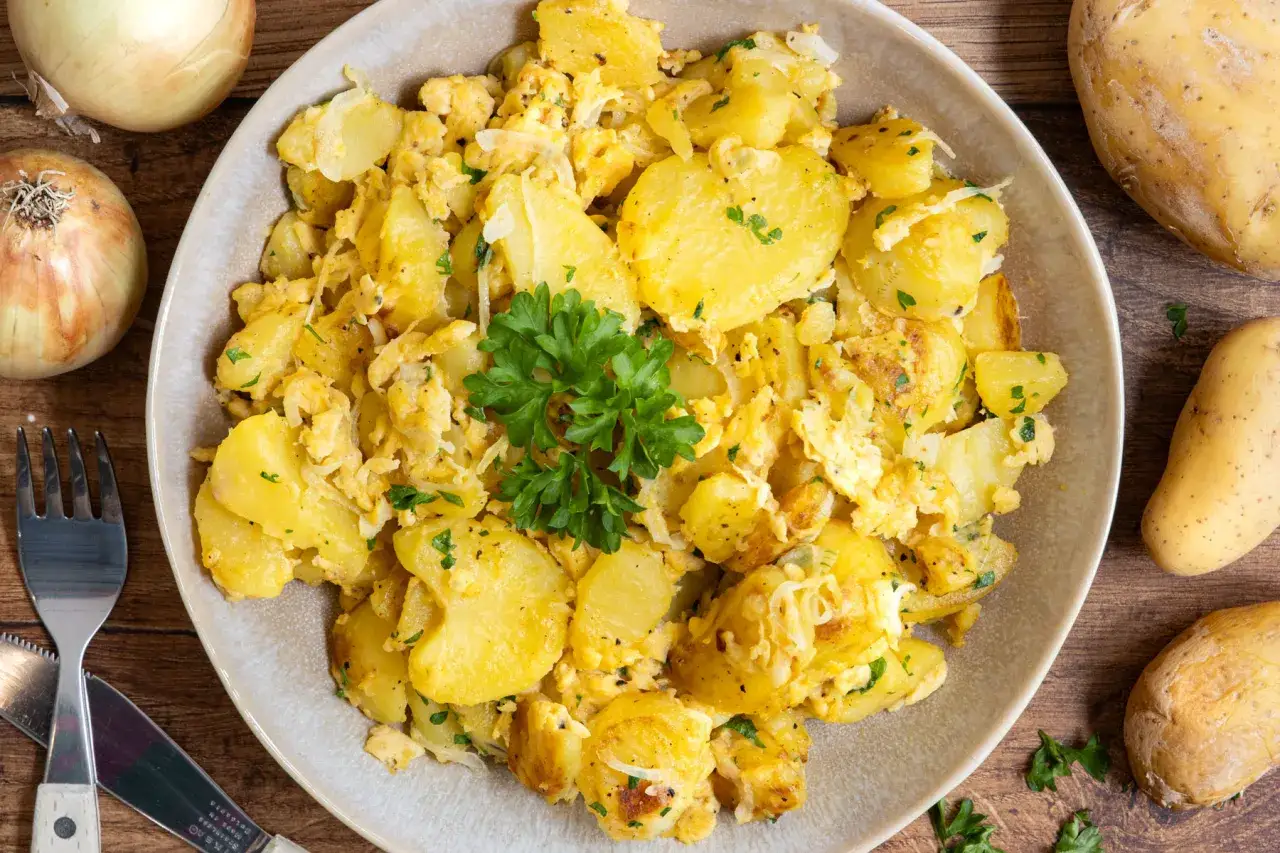 Herzhafte bratkartoffeln mit ei, garniert mit Petersilie, auf einem Teller. Daneben liegen rohe Kartoffeln und Zwiebeln.