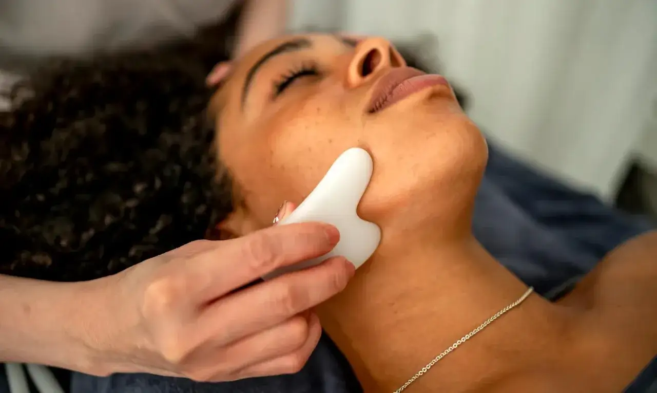 A white gua sha stone is used to perform face massage steps on a woman's jawline, promoting relaxation and skin rejuvenation.