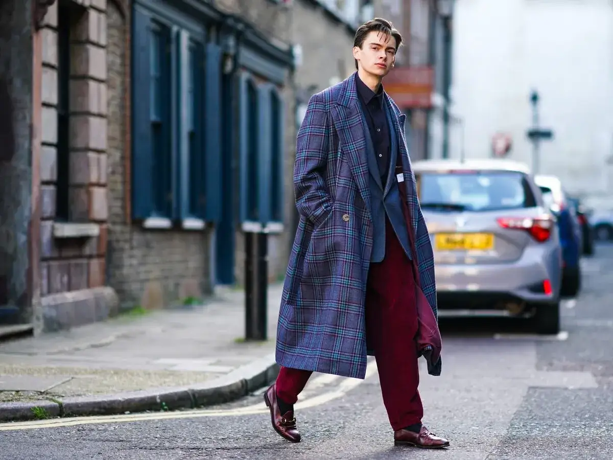 Un uomo con un cappotto a quadri, un completo grigio e pantaloni bordeaux cammina per strada, mostrando diversi tipi di cappotto.