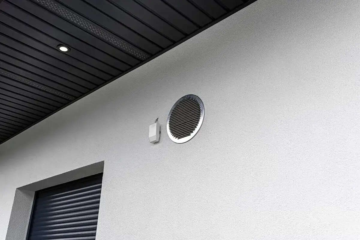 Grille de ventilation ronde sur un mur blanc, avec un petit boîtier blanc à côté. Un élément essentiel pour la ventilation de votre appartement.