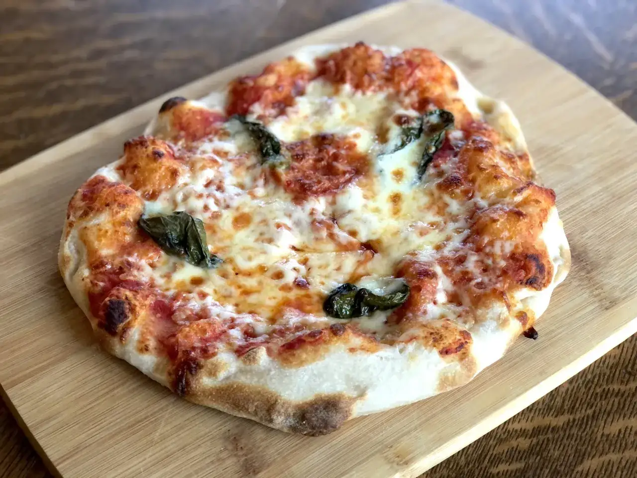 Eine kleine Pizza mit geschmolzenem K&auml;se und Basilikum auf einem Holzbrett. Der Teig, der 24 Stunden gereift ist, hat eine luftige Kruste.