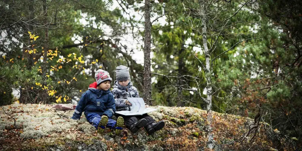 Två barn njuter av aktiviteter i skogen med barn, sitter på en mossig sten och läser en bok bland höstfärgade träd.