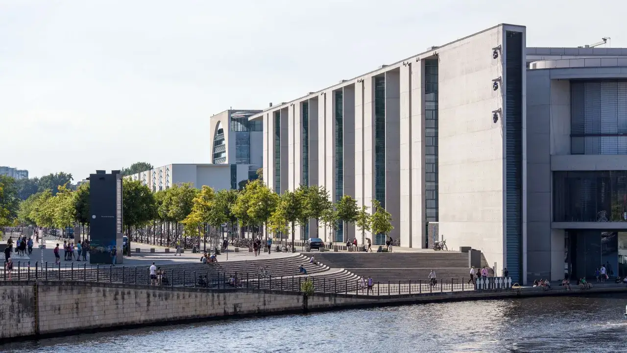 Moderne Architektur am Spreeufer in Berlin, die an die Grenzen der Macht des Bundeskanzlers erinnert. Menschen flanieren, sitzen und genie&szlig;en die Sonne.