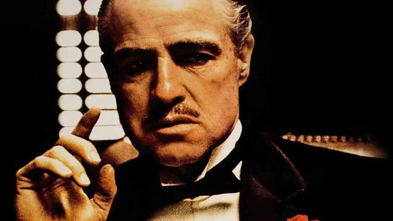 Marlon Brando jako ojciec chrzestny, z charakterystycznym wąsem i gestem dłoni, w mrocznym wnętrzu.