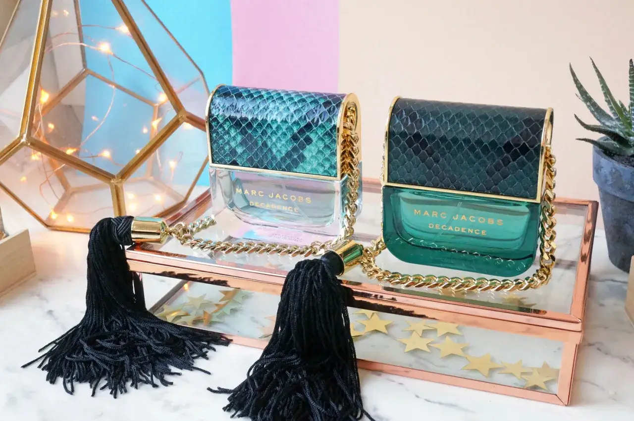 Dwa flakoniki perfum Marc Jacobs Decadence Eau So Decadent w zielonych, wężowych opakowaniach, ozdobione złotymi łańcuszkami i czarnymi chwostami.