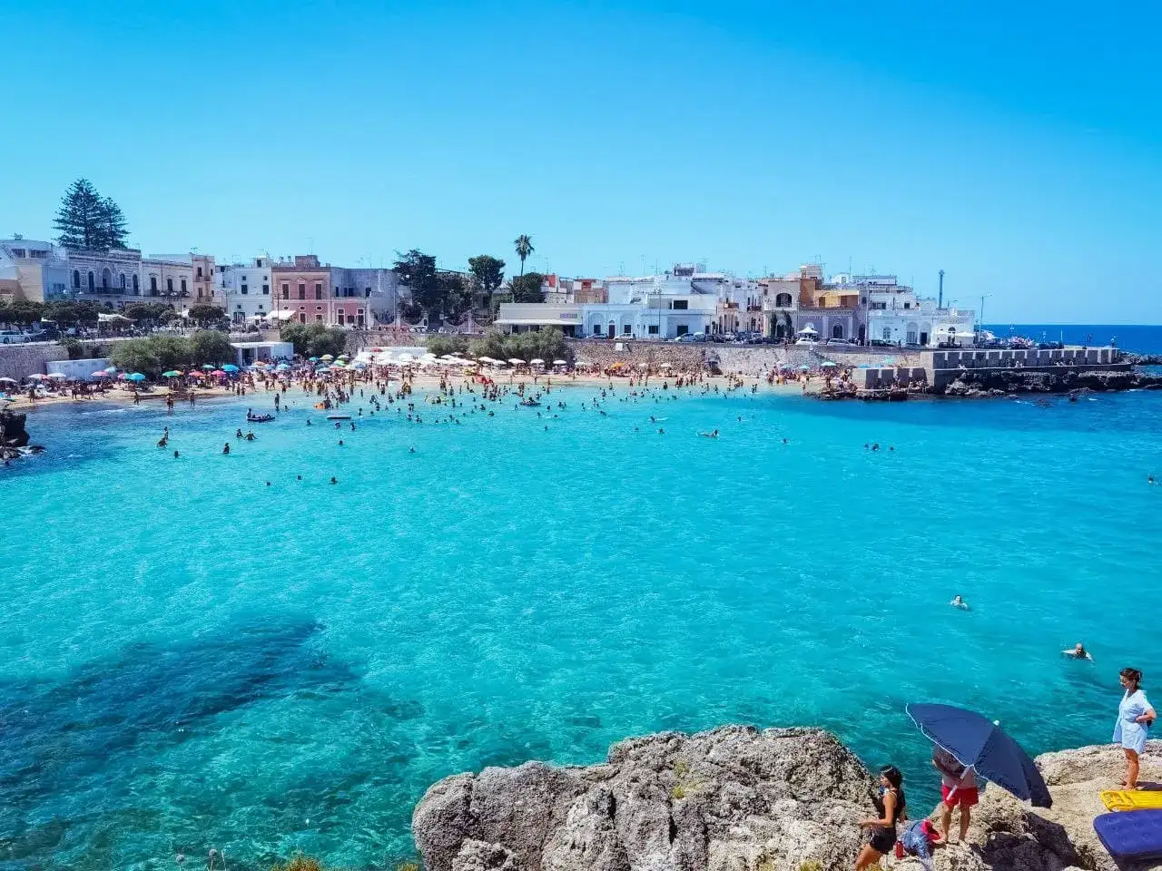 Una spiaggia affollata con acque cristalline, bagnanti e case bianche sullo sfondo. Un luogo da bandiera blu, perfetto per una vacanza estiva.