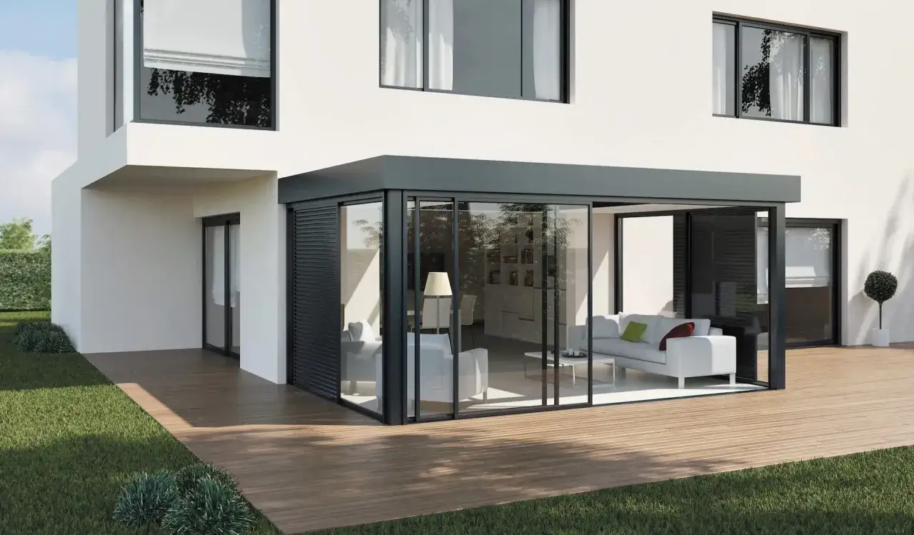 Une veranda moderne, conçue par un architecte, s'ouvre sur une terrasse en bois. L'intérieur est lumineux avec un canapé blanc.