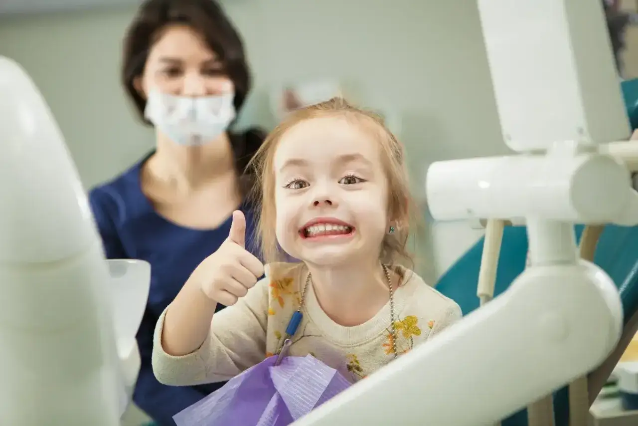 Uśmiechnięta dziewczynka pokazuje kciuk w górę u dentysty, gdy wypadają jej jedynki górne.