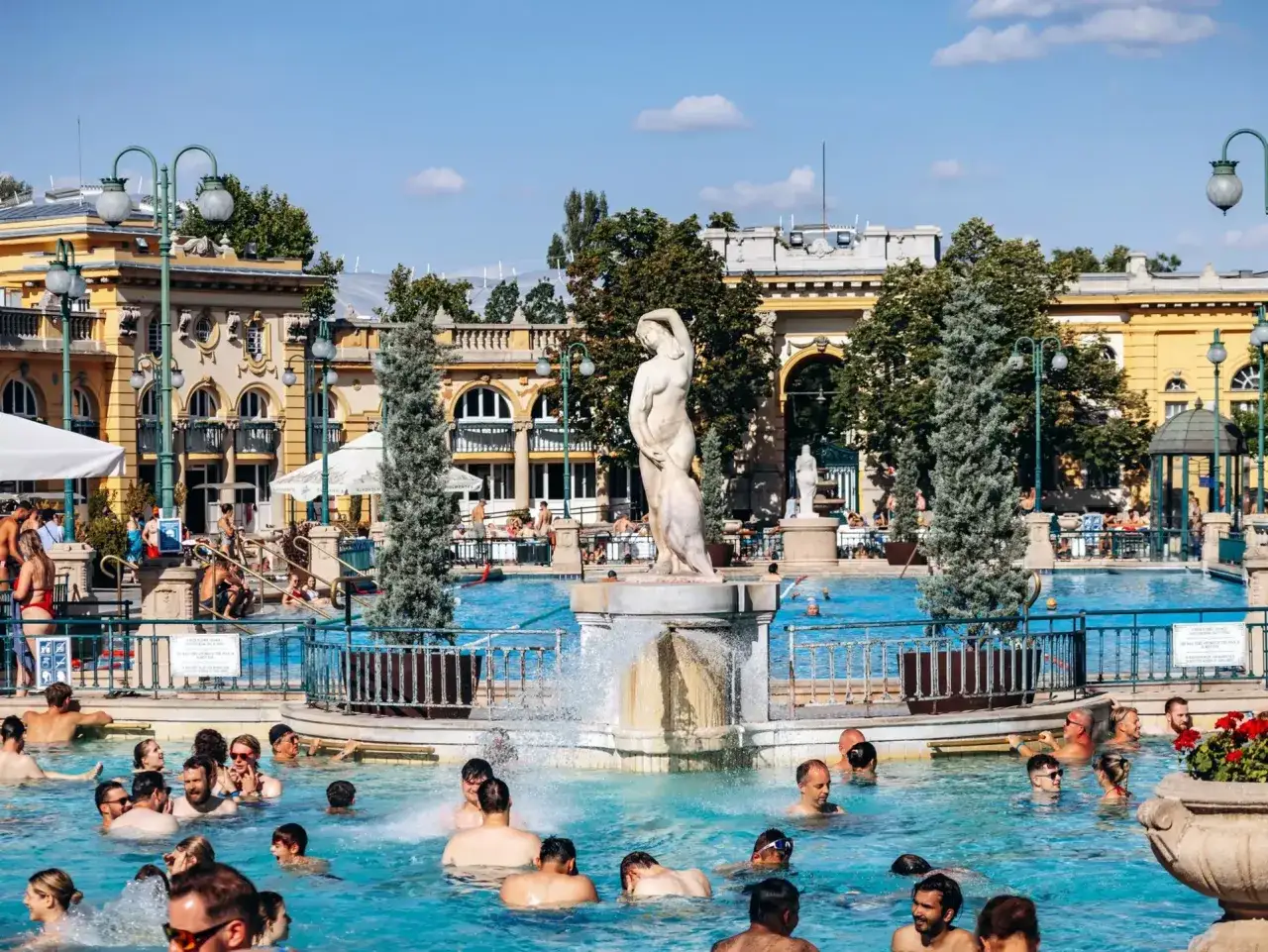 M&auml;nniskor njuter av ett termalbad i Budapest, med en staty och gul byggnad i bakgrunden.