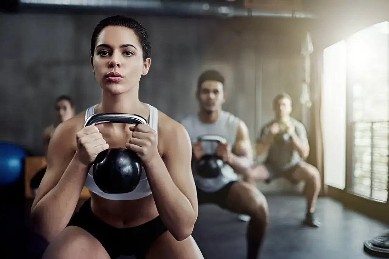 Kobieta wykonuje przysiady z kettlebell, skupiona na ćwiczeniach siłowych. W tle inni ćwiczą.