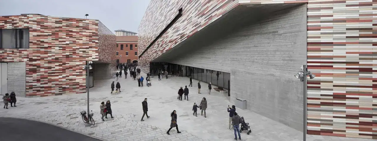 Esterno moderno del museo del Novecento a Mestre, con facciate colorate e cemento a vista. Persone passeggiano nella piazza antistante.