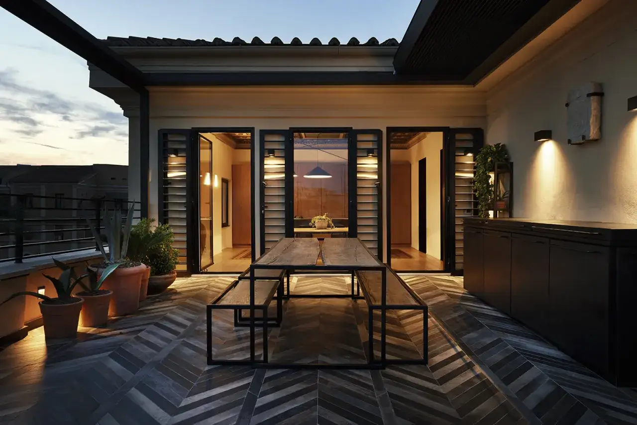 Terrazza con tavolo in legno e panche, illuminata da luci soffuse. La stanza buia all'interno &egrave; visibile attraverso le vetrate.