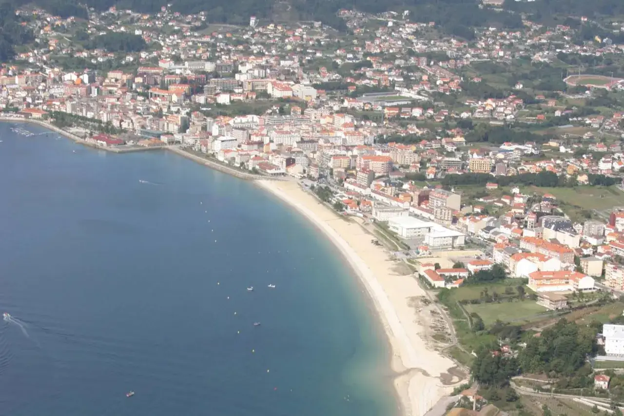 Vista aérea de la **playa de Rodeira**, con el mar azul, la arena dorada y la ciudad extendiéndose hasta las colinas verdes.