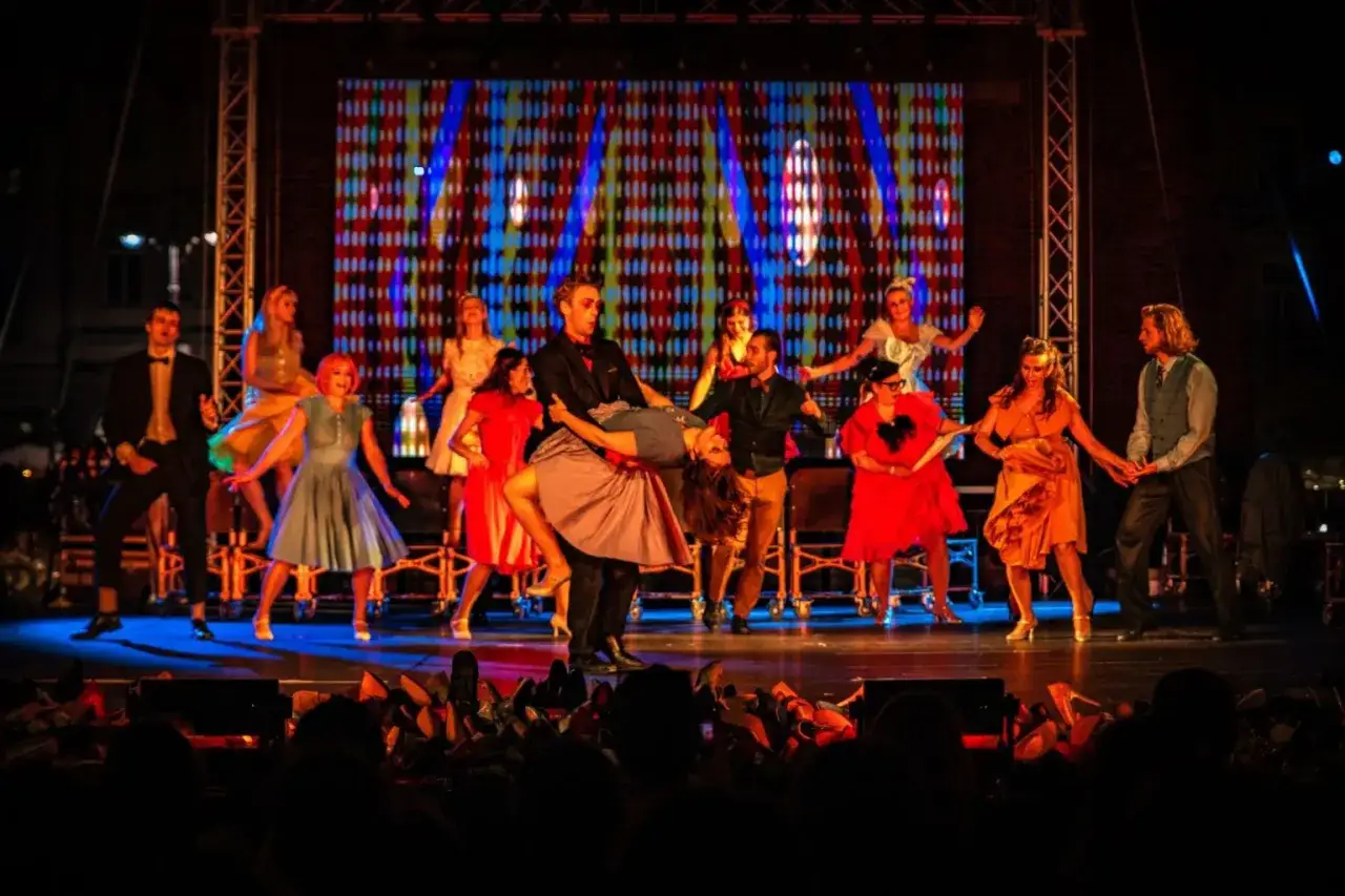 Tancerze w barwnych strojach wykonują dynamiczny spektakl w teatrze. W tle migocze kolorowa iluminacja.