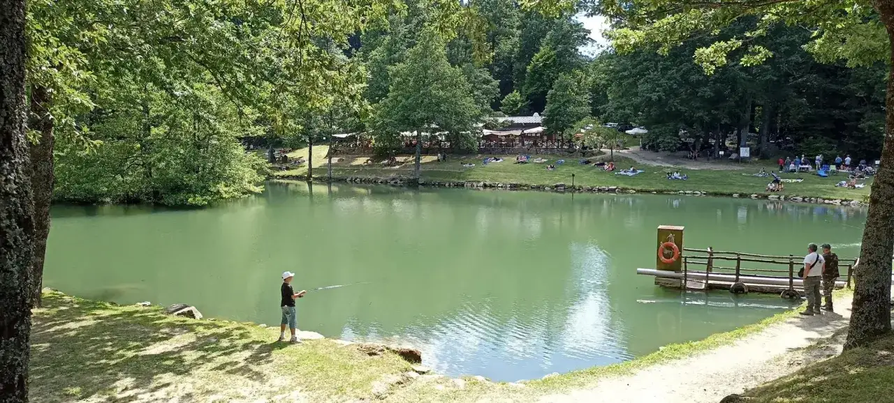 Un ragazzo pesca nel lago Pontini, circondato da alberi e persone che si rilassano sull'erba.