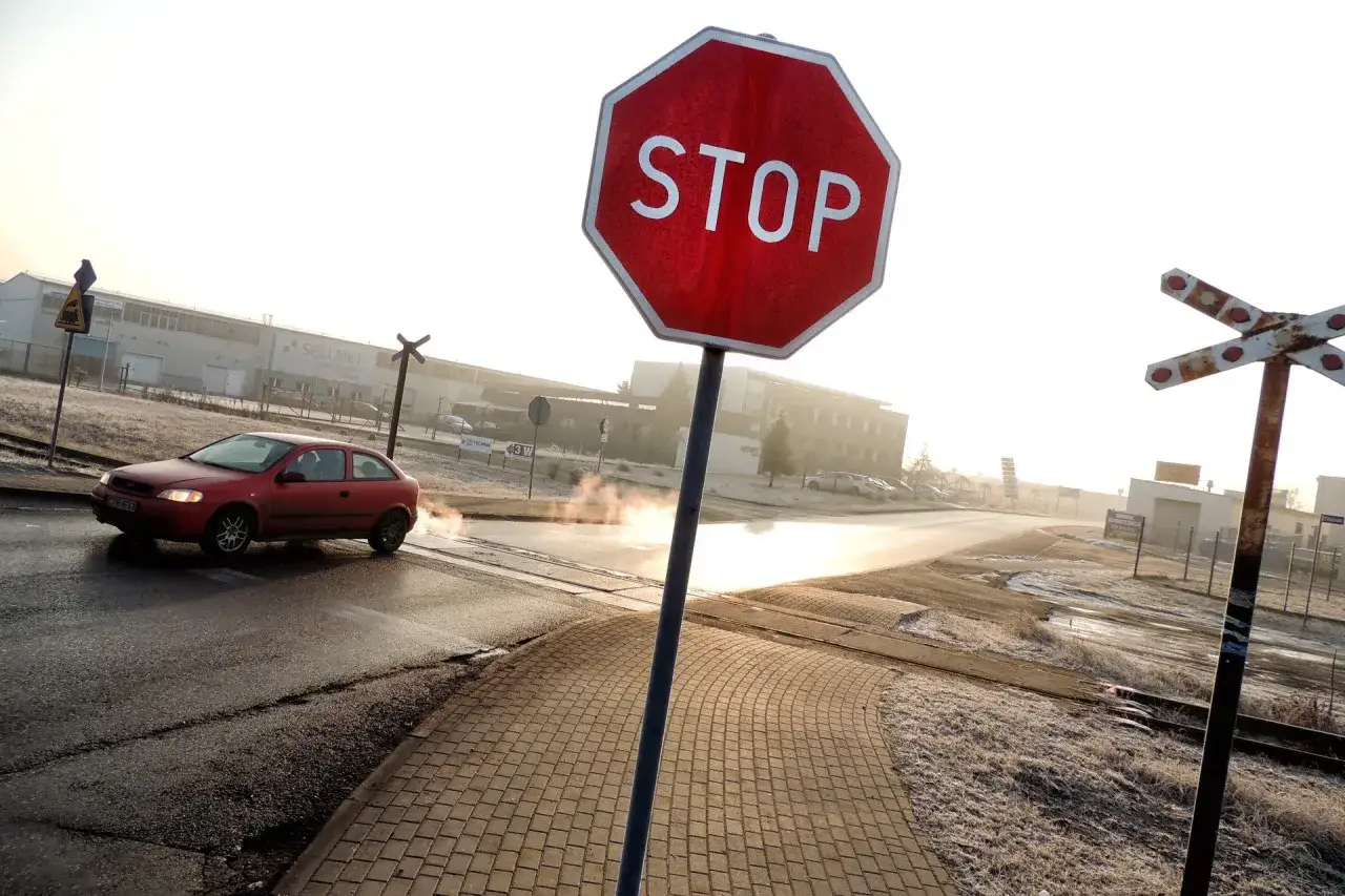 Czerwony samochód zbliża się do przejazdu kolejowego, ignorując znak STOP. Para unosi się z mokrej drogi.