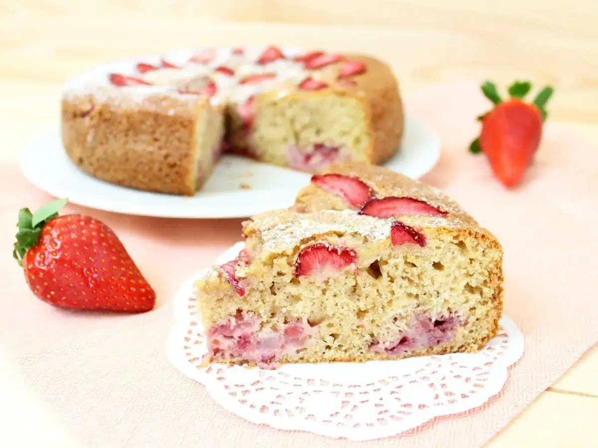 Una fetta di torta di fragole e yogurt, cosparsa di zucchero a velo, con fragole fresche e la torta intera sullo sfondo.