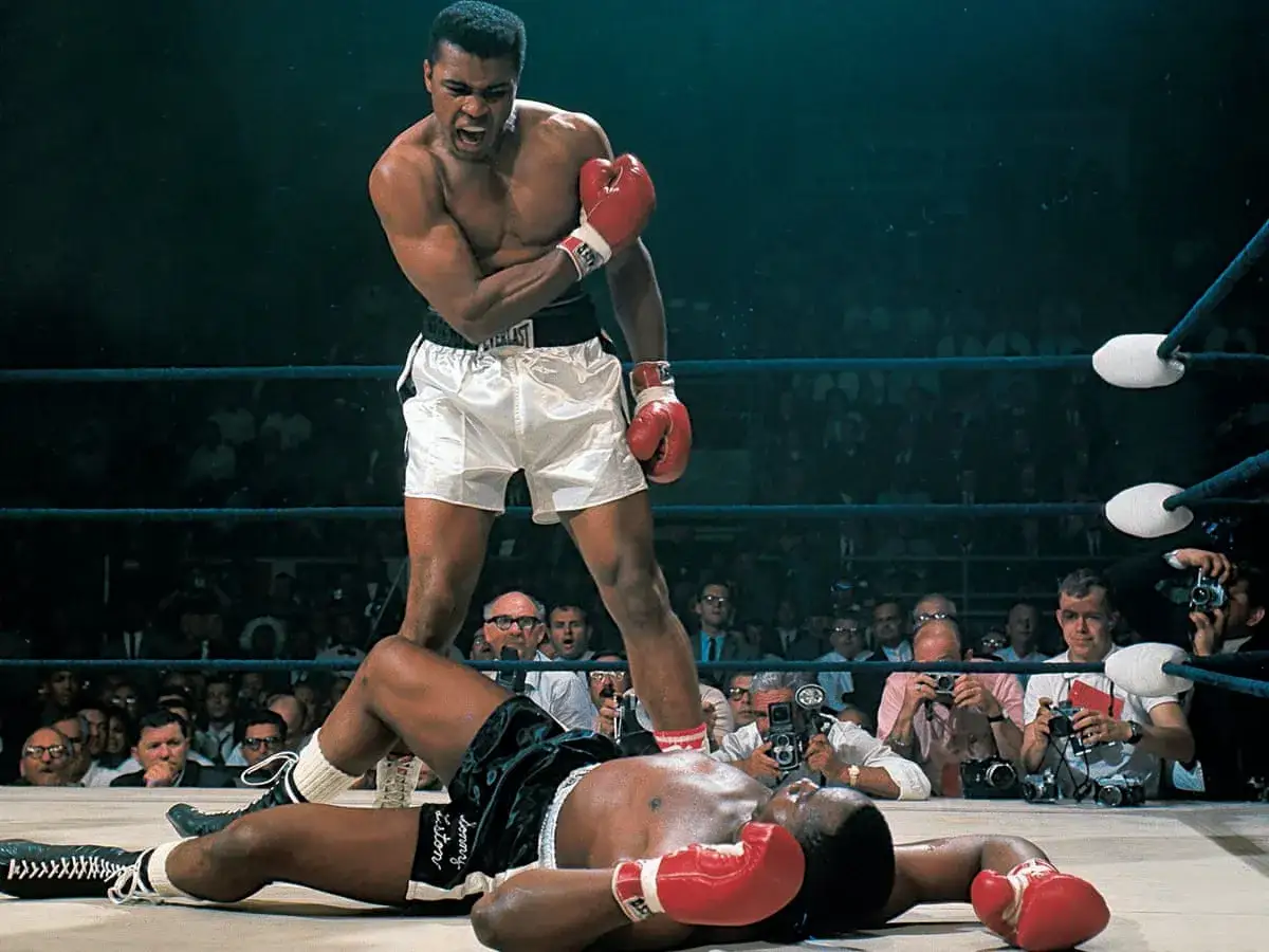 Muhammad Ali trionfa sul ring, un momento iconico catturato da fotografi sportivi famosi. Un pugile a terra, l'altro in piedi, esultante.