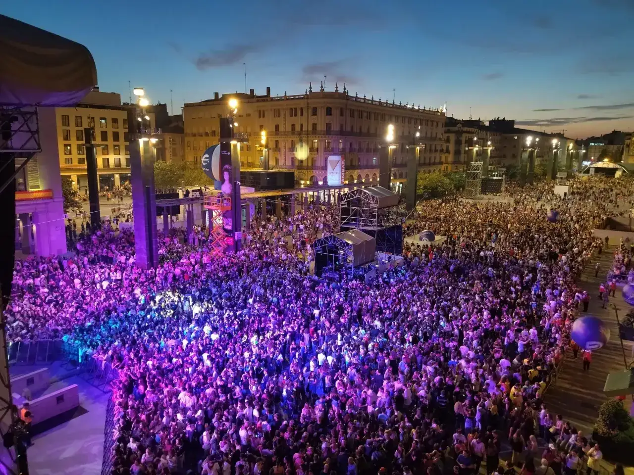 Multitud disfrutando de un concierto de música al raso, iluminado por luces moradas y azules.
