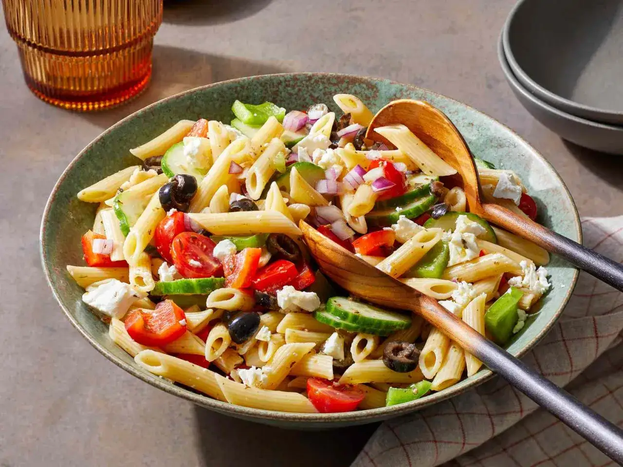 En stor skål med grekisk pastasallad med penne, tomat, gurka, paprika, rödlök, oliver och fetaost. Serveras med träskedar.