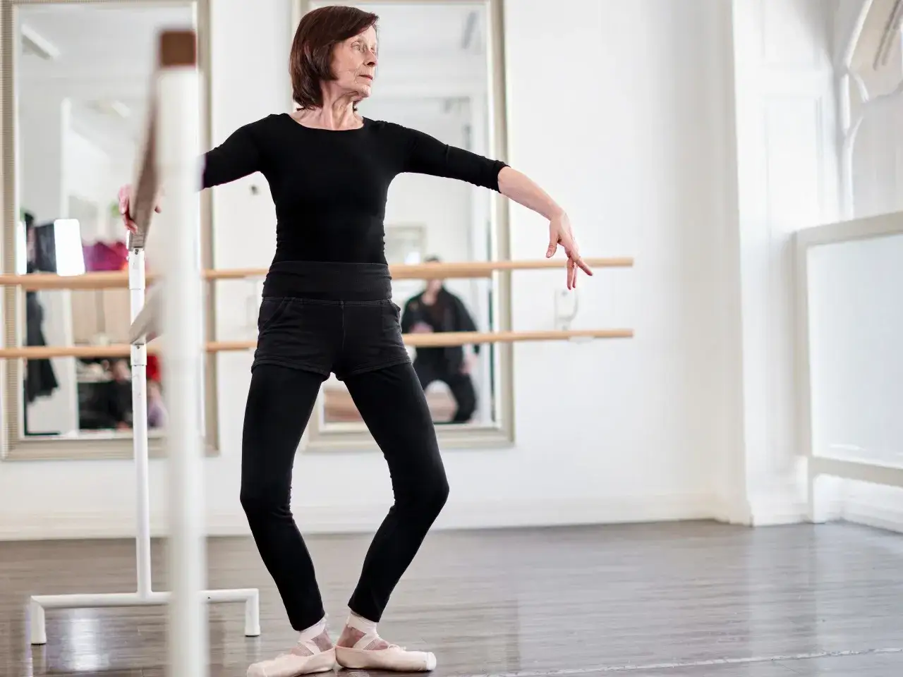 Ältere Frau in schwarzer Kleidung und Ballettschuhen übt Barre Training an einer Ballettstange.