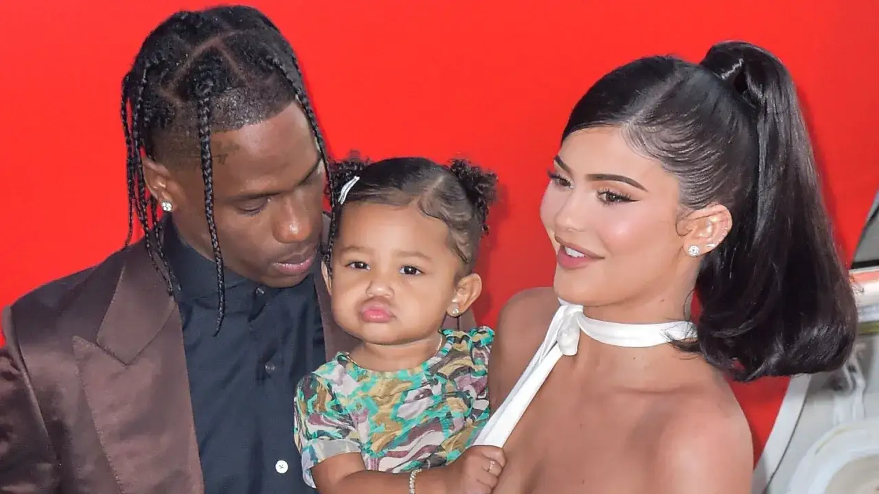 Kylie Jenner z dzieckiem i Travisem Scottem na czerwonym tle.