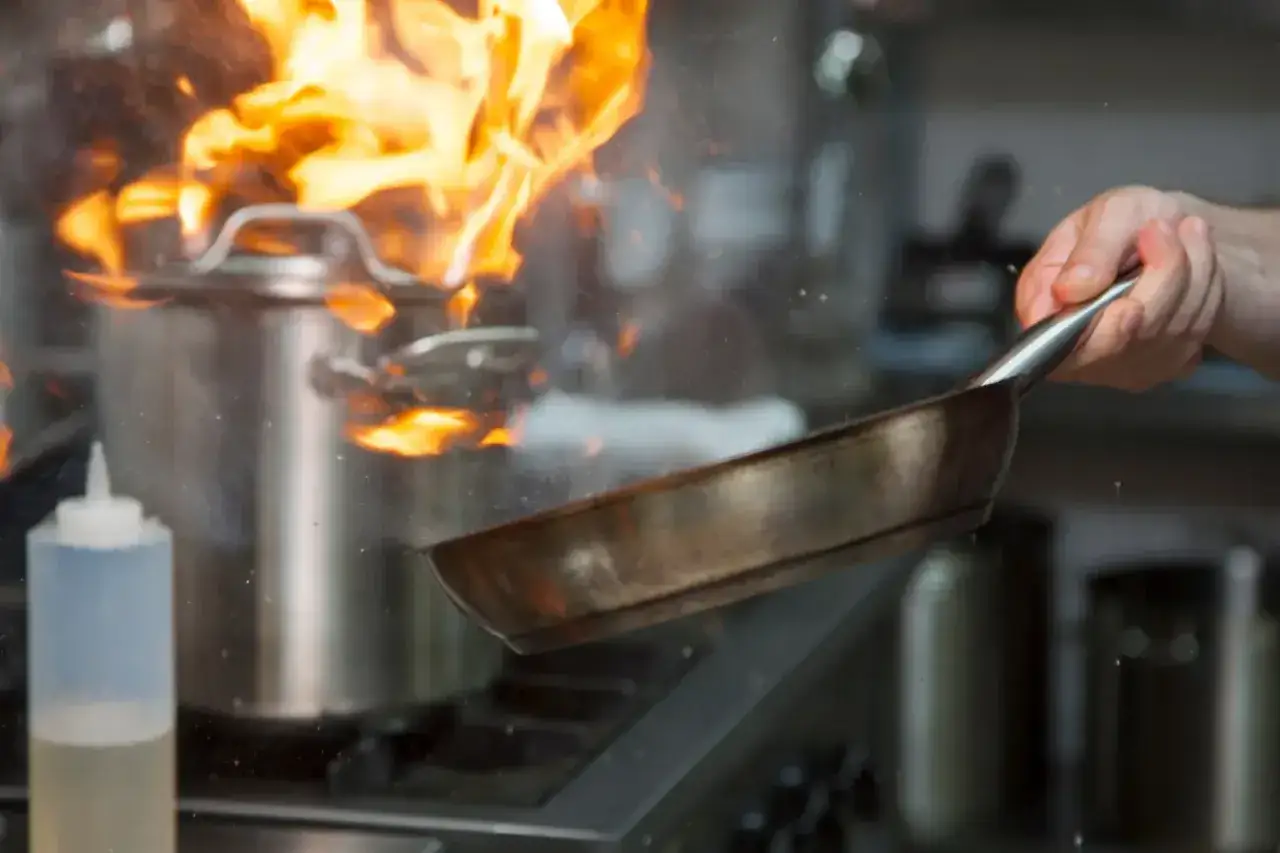 Mistrz kuchni wykonuje flambirowanie, unosząc patelnię nad płomieniami buchającymi z garnka.