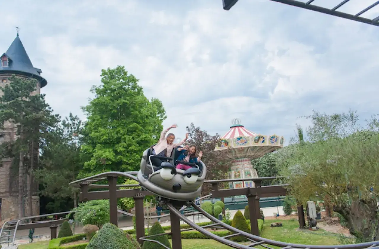 Deux enfants s'amusent dans un wagon de montagnes russes en forme de chenille dans un parc d'attractions. Un carrousel et un château complètent le décor.