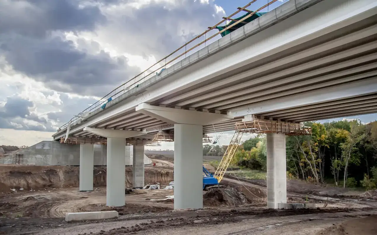Construcción de puentes de acero con pilares de hormigón. Una grúa amarilla y azul trabaja en el sitio.