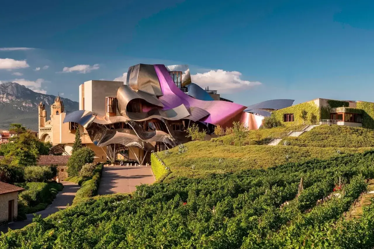 Edificio vanguardista con tejado metálico púrpura y plateado, rodeado de viñedos y vegetación. Un ejemplo de alojamientos originales España.