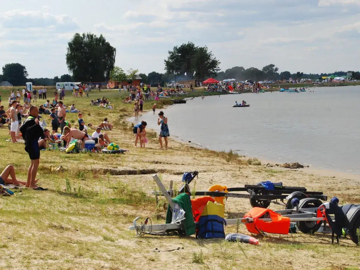 Ludzie wypoczywają nad wodą, na plaży i w wodzie. Widać łodzie, kamizelki ratunkowe i przyczepę. To dzień na zalewie biszcza.
