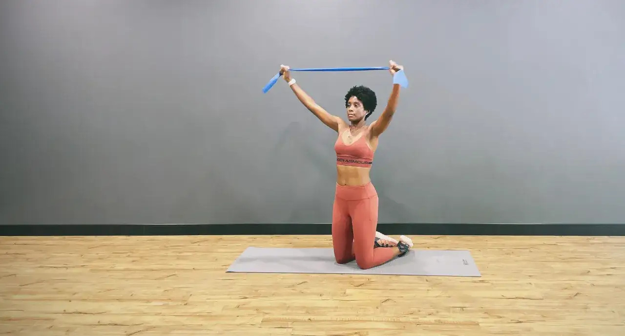 Femme à genoux sur un tapis, effectuant un exercice haut du corps avec une bande élastique bleue au-dessus de sa tête.