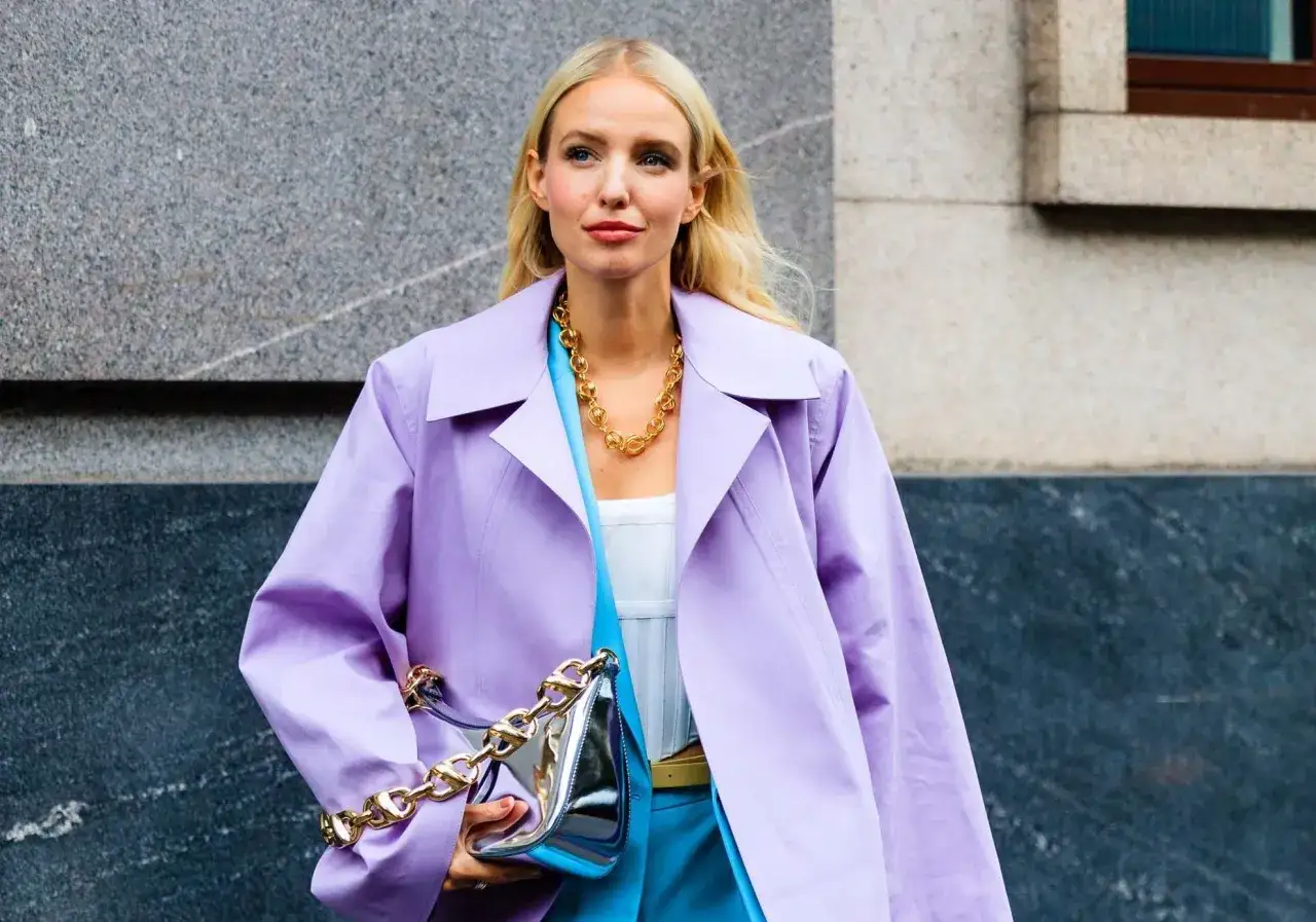 Une femme arbore une tenue mi saison femme tendance : manteau lavande, top blanc, pantalon bleu vif, sac argenté et collier doré.