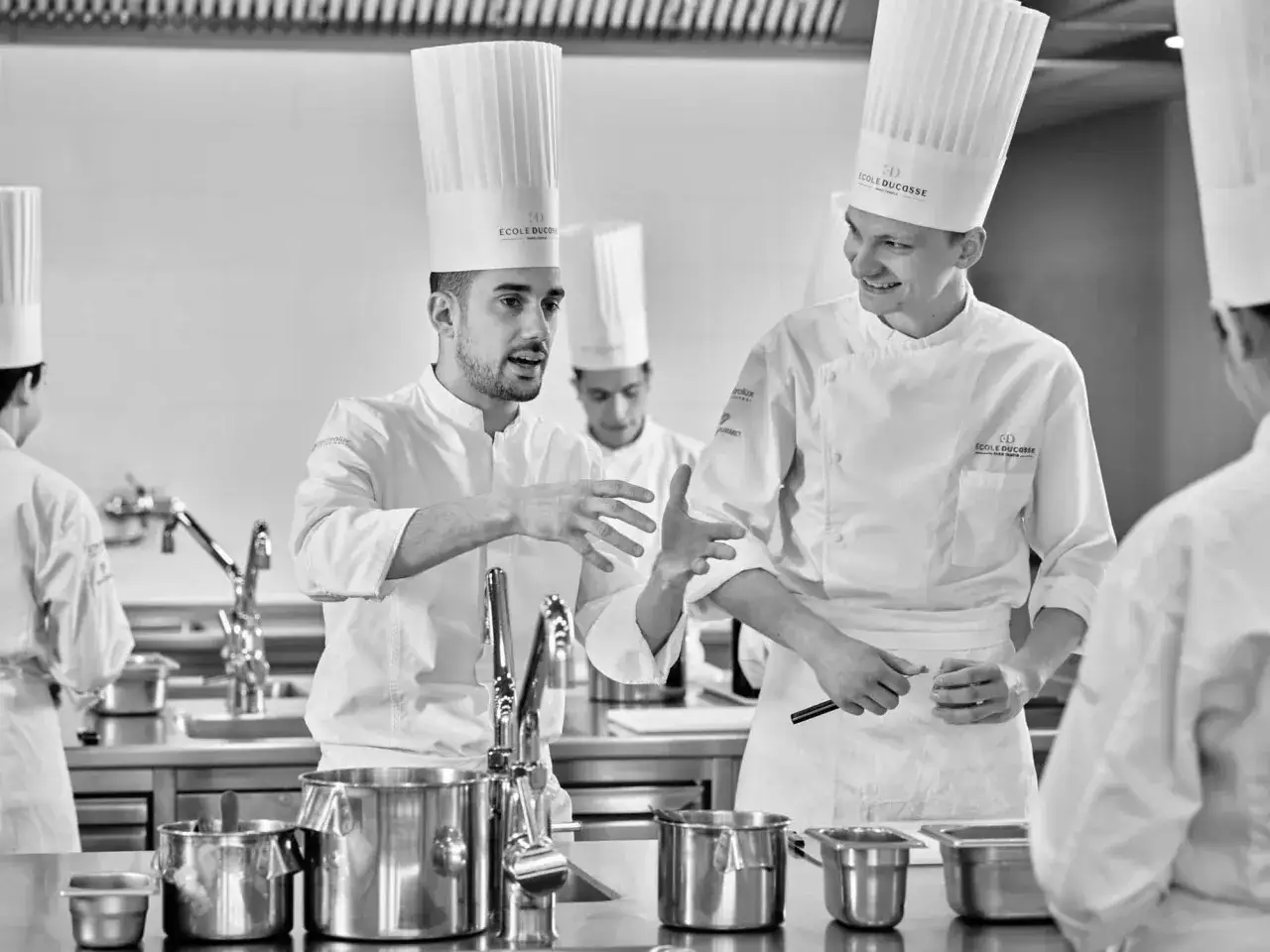 Dos chefs con gorros altos discuten técnicas culinarias, uno gesticula apasionadamente. Parecen estar en camino de convertirse en el mejor cocinero del mundo.
