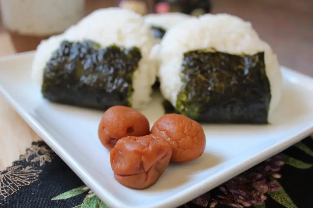 Two onigiri wrapped in nori sit on a white plate, with three pickled plums in front. A simple, classic hinomaru bento.