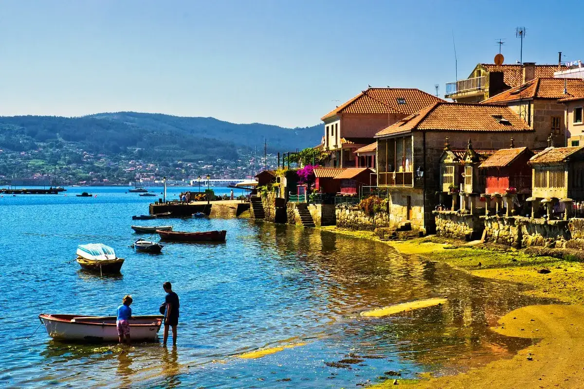 Dos niños en una barca observan la costa de uno de los pueblos bonitos de Galicia, con casas de piedra y tejados rojos junto al mar azul.