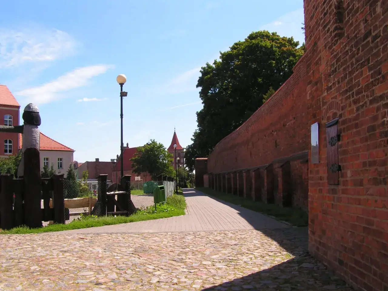 Spacer po historycznej trasie w Chełmnie. Brukowana ścieżka wzdłuż ceglanych murów obronnych, z widokiem na kamienice i wieżę kościelną.