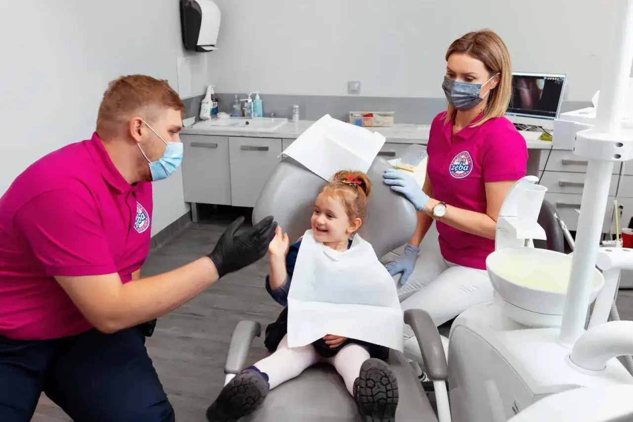 Uśmiechnięta dziewczynka w gabinecie dentystycznym, otoczona przez personel. Bezpieczna sedacja minimalizuje skutki uboczne.