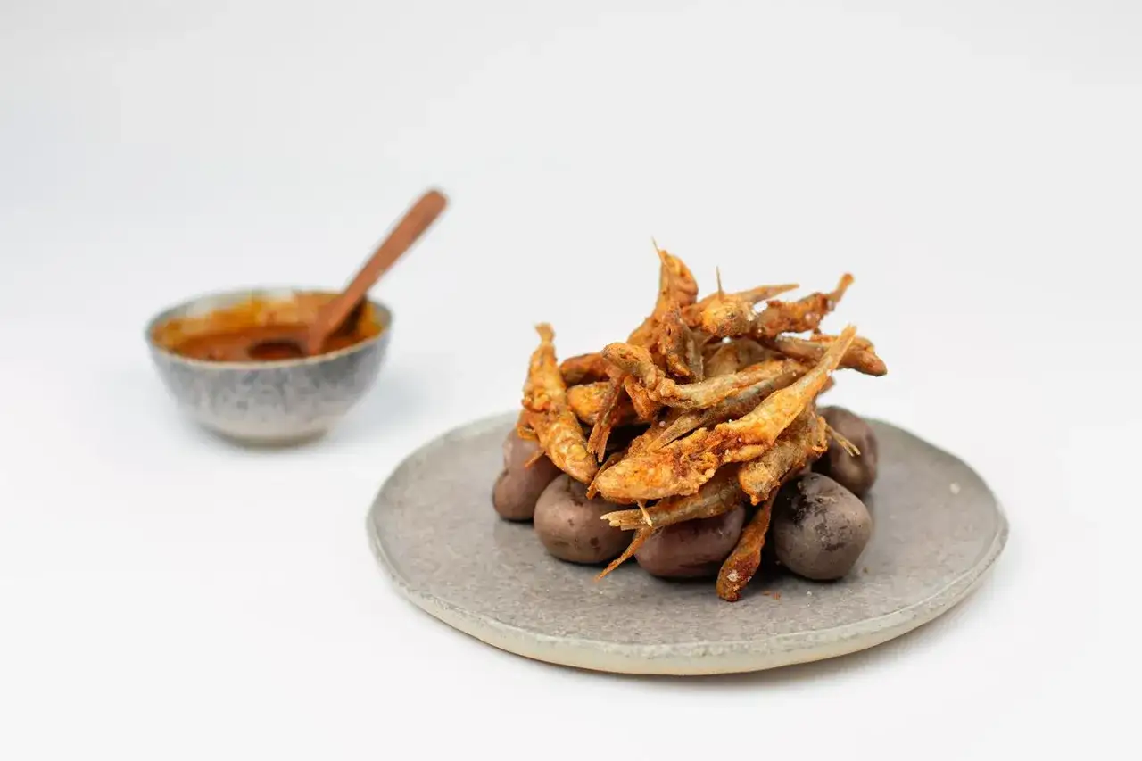 Un tas de petits poissons croustillants, l'**éperlan frit**, repose sur des pommes de terre violettes. Un bol de sauce épicée est à gauche.