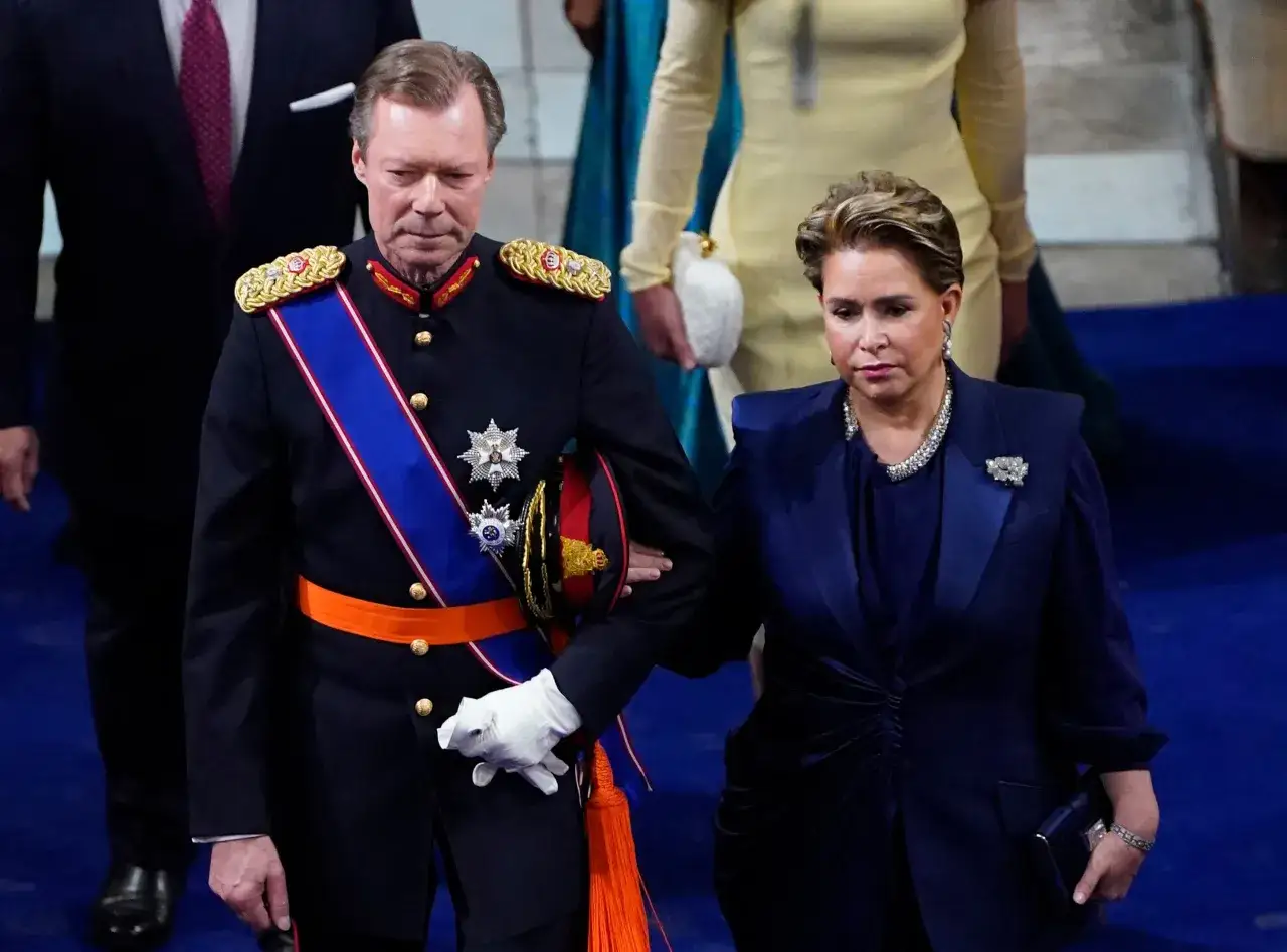 Henri av Luxemburg i milit&auml;runiform med medaljer och en bl&aring; och orange banderoll. Bredvid honom g&aring;r en kvinna i m&ouml;rkbl&aring; kl&auml;nning.