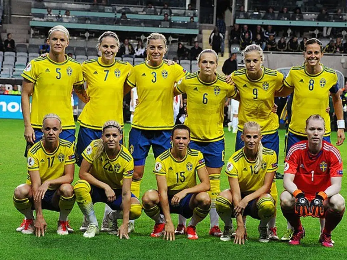Svenska kvinnliga fotbollsspelare i gula tröjor står och sitter på en gräsplan.