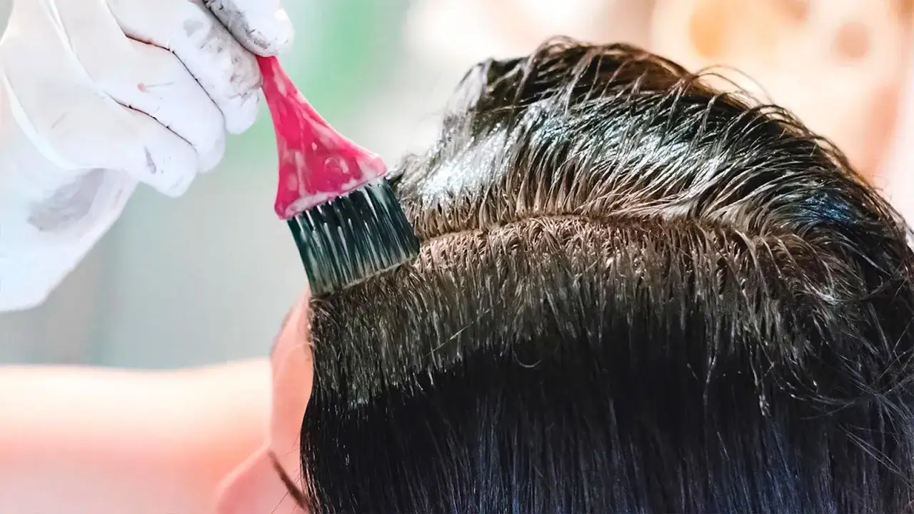 Aplicando tinte para te&ntilde;ir pelo con canas en casa. Guantes blancos y brocha rosa cubren las ra&iacute;ces oscuras.