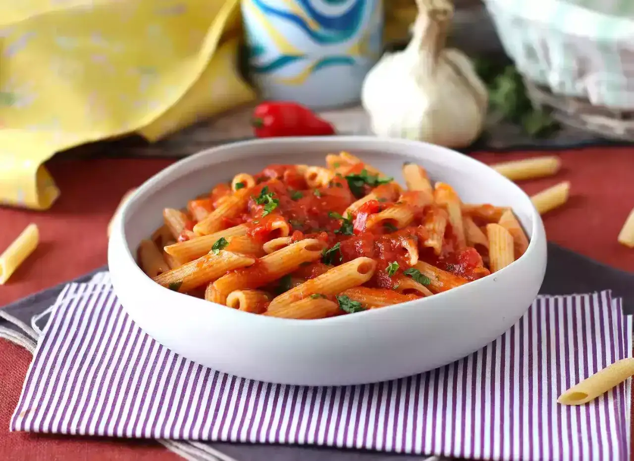 Penne arrabiata w białej misce, ozdobione natką pietruszki. Obok czosnek i papryczka chili.