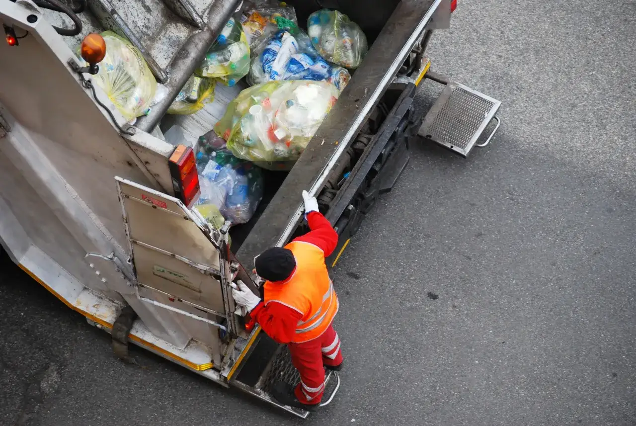 Śmieciarka z workami pełnymi odpad&oacute;w. Pracownik w kamizelce odblaskowej i czerwonym stroju przygotowuje się do załadunku. Zastanawiasz się, ile kosztuje tona śmieci na wysypisku?