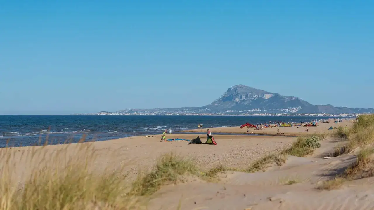 Gente disfrutando de la playa de Oliva, con kitesurfistas y tiendas en la arena. Montaña al fondo. Opiniones positivas para este destino.