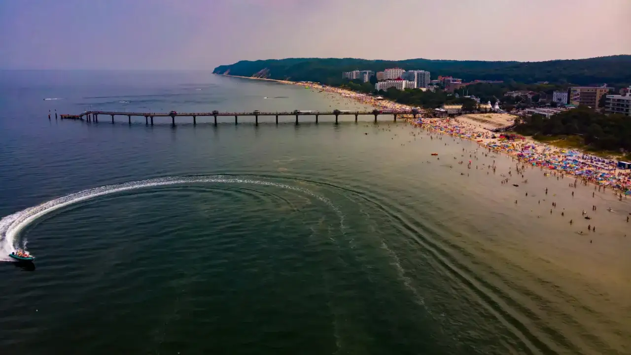 Ein belebter Strand in Misdroy, Polen, mit einer langen Seebrücke und einem Schnellboot, das eine Spur im Wasser hinterlässt.
