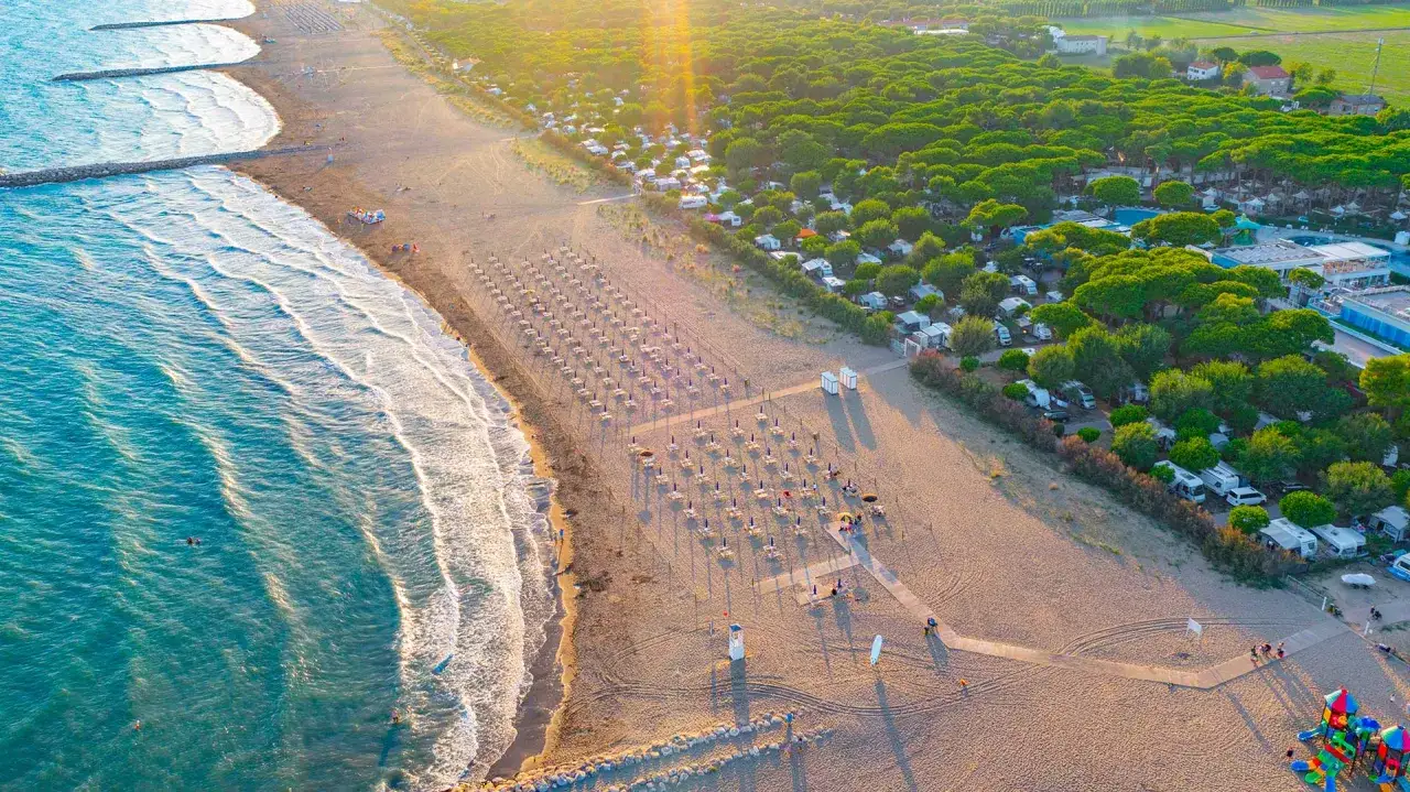 Widok z lotu ptaka na plażę z rzędami leżaków i parasoli, a za nią rozciąga się camping residence village z kamperami i zielonymi drzewami.