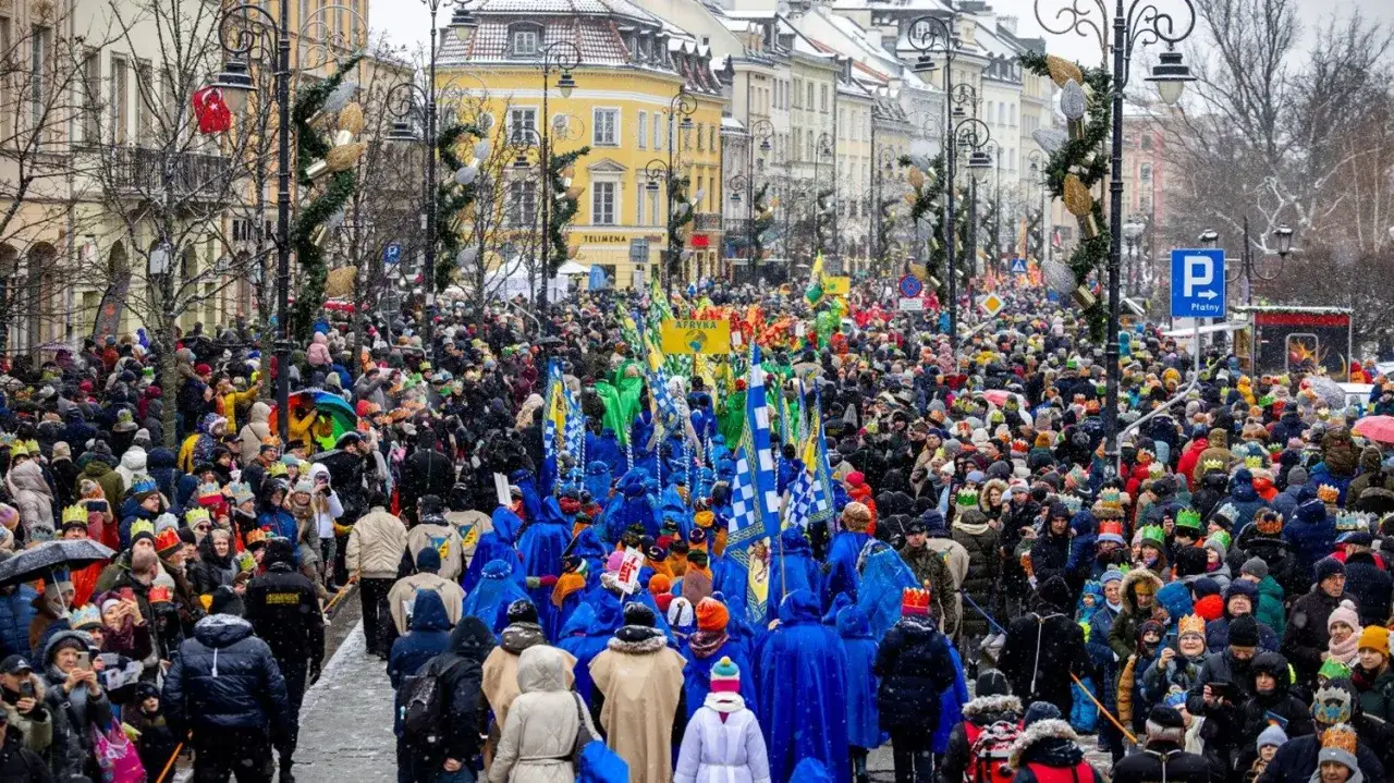 Tłum ludzi w koronach na głowach, maszeruje ulicą podczas Orszaku Trzech Króli. Wszędzie flagi i kolorowe stroje.